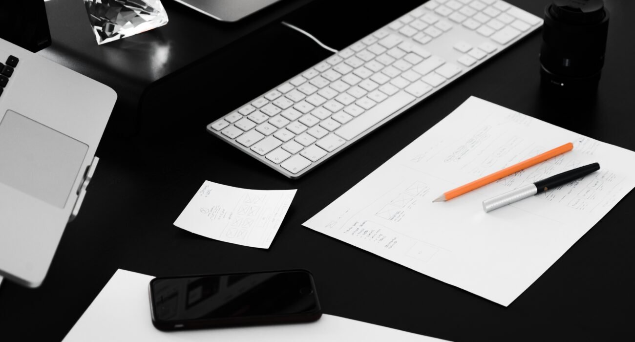 Web designer's workspace featuring a laptop, keyboard, design sketches, smartphone, and creative planning materials on a desk.