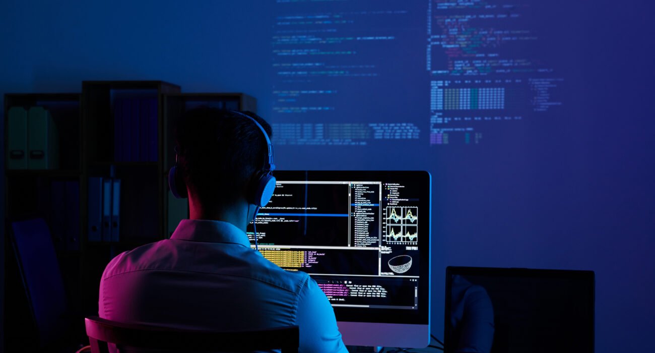Software developer working on code at a computer workstation with multiple programming screens in a dark office.