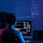 Software developer working on code at a computer workstation with multiple programming screens in a dark office.