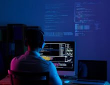 Software developer working on code at a computer workstation with multiple programming screens in a dark office.